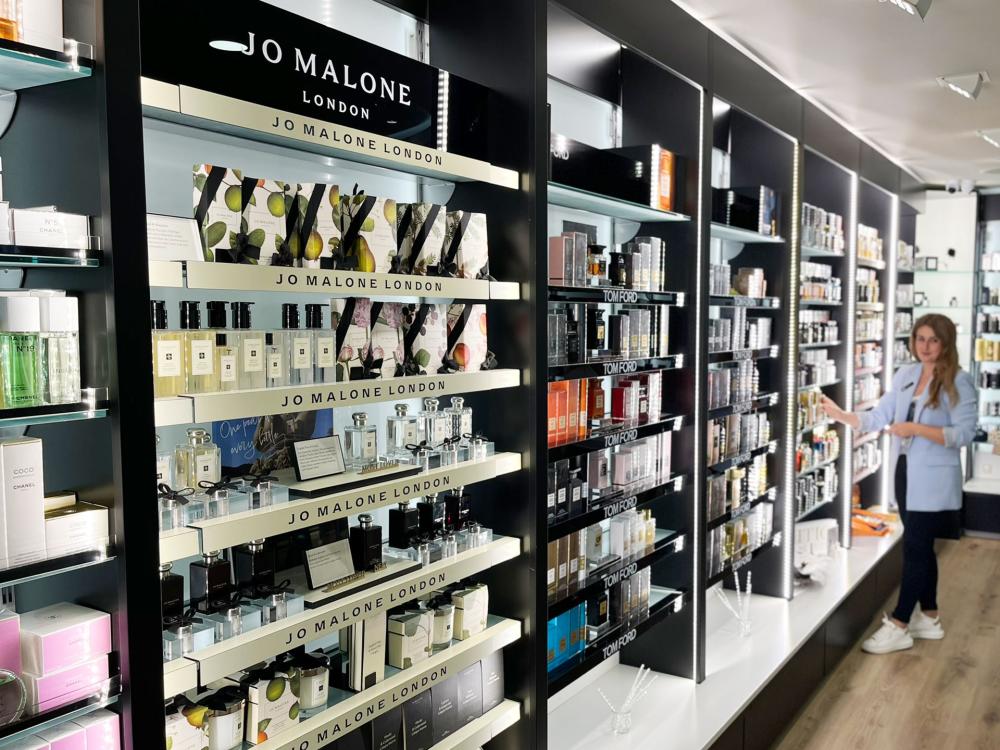 Innenaufnahme einer modernen Parfümerie mit beleuchteten Regalen. Im Vordergrund stehen Düfte und Pflegeprodukte der Marke Jo Malone London, daneben Parfums von Tom Ford. Rechts im Bild steht eine Frau in hellblauer Jacke vor den Regalen und betrachtet die Produkte. Die Atmosphäre wirkt elegant und hochwertig.
