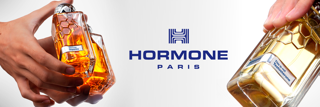 Zwei Hände halten futuristisch geformte Parfumflakons in warmen Gold- und Bernsteintönen vor hellem Hintergrund. In der Mitte steht das blaue Logo mit dem Schriftzug „HORMONE PARIS“. Auf den Etiketten der Flakons stehen Hinweise wie „This is not Dopamine“