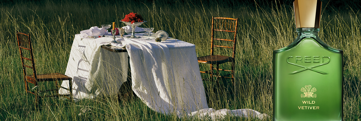 In einer natürlichen Wiesenlandschaft steht ein gedeckter Tisch mit weißer Tischdecke, Blumen und Geschirr, daneben ein leerer Stuhl. Rechts im Vordergrund ist ein grüner Parfumflakon mit goldener Kappe zu sehen. Im Hintergrund erkennt man hohes Gras und 