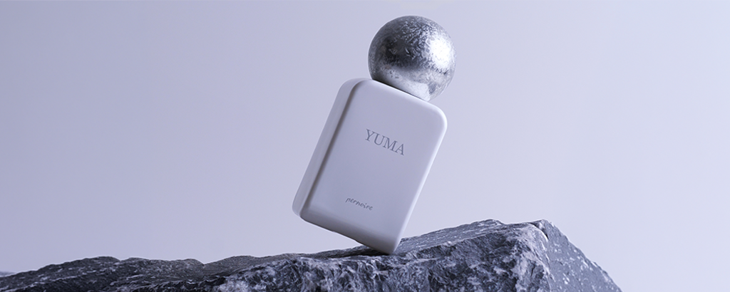 Minimalistisches Duftbild: Weißer Parfumflakon mit der Aufschrift ‚Yuma‘ und kugelförmigem silbernen Verschluss steht schräg auf dunklem Felsen. Kühle, graublaue Lichtstimmung vor schlichtem Hintergrund.