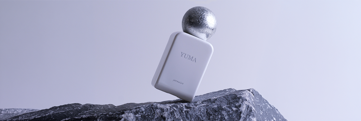 Minimalistisches Duftbild: Weißer Parfumflakon mit der Aufschrift ‚Yuma‘ und kugelförmigem silbernen Verschluss steht schräg auf dunklem Felsen. Kühle, graublaue Lichtstimmung vor schlichtem Hintergrund.