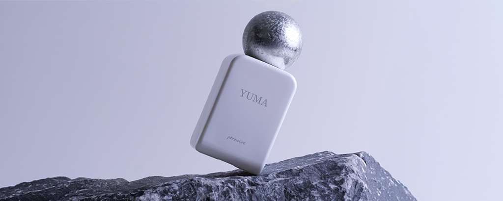 Minimalistisches Duftbild: Weißer Parfumflakon mit der Aufschrift ‚Yuma‘ und kugelförmigem silbernen Verschluss steht schräg auf dunklem Felsen. Kühle, graublaue Lichtstimmung vor schlichtem Hintergrund.
