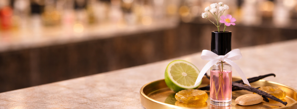 Dekorative Duftabfüllung auf goldenem Tablett: kleines Glasfläschchen mit rosafarbener Flüssigkeit, schwarzem Deckel und weißer Schleife, verziert mit zarten Blüten. Daneben liegen eine halbe Limette, Vanilleschoten und Harzstücke. Unscharfer Parfümregal-Hintergrund.