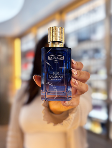 Blauer Glasflakon des Parfums Blue Talisman von Ex Nihilo mit goldenem Verschluss, in einer Hand gehalten vor unscharfem Parfümerieregal.