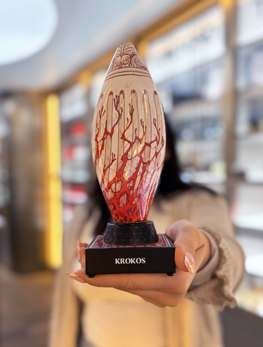 Skulpturaler Parfumflakon Krokos mit cremefarbener Oberfläche und roten, korallenartigen Mustern auf schwarzem Sockel, in einer Hand vor Parfümeriehintergrund gehalten.