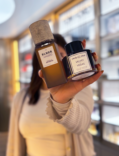 Hand hält zwei Nischenparfumflakons vor einem Parfümerieregal: ein brauner Flakon mit Naturkork-Verschluss und ein dunkler Flakon mit schwarzem Deckel, beide mit minimalistischen Etiketten.