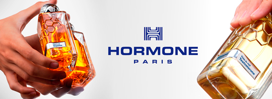 Zwei Hände halten futuristisch geformte Parfumflakons in warmen Gold- und Bernsteintönen vor hellem Hintergrund. In der Mitte steht das blaue Logo mit dem Schriftzug „HORMONE PARIS“. Auf den Etiketten der Flakons stehen Hinweise wie „This is not Dopamine“ und „This feels like Testosterone“