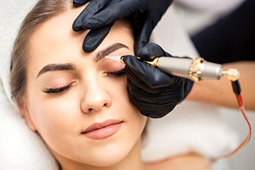 Frau mit geschlossenen Augen erhält ein Permanent-Make-up an den Augenbrauen von einer Kosmetikerin mit schwarzen Handschuhen und speziellem Gerät.