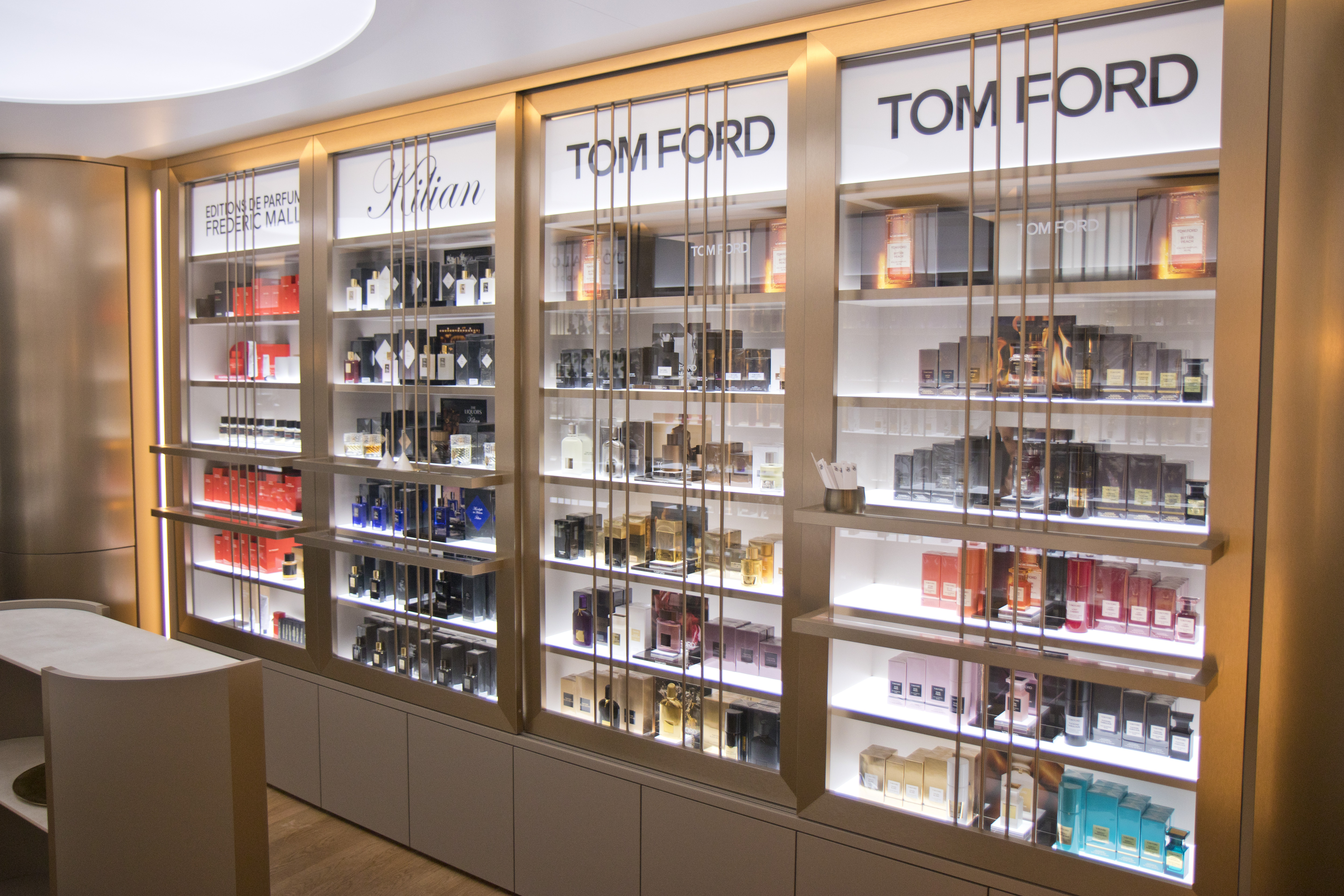 Beleuchtete Regalwand in einer Parfümerie mit hochwertigen Düften von Marken wie Editions de Parfums Frédéric Malle, Kilian und Tom Ford. Die Parfums sind in elegant gestalteten Flakons und Verpackungen arrangiert, die Regale sind in warmem Goldrahmen gehalten und schaffen eine luxuriöse, moderne Atmosphäre.