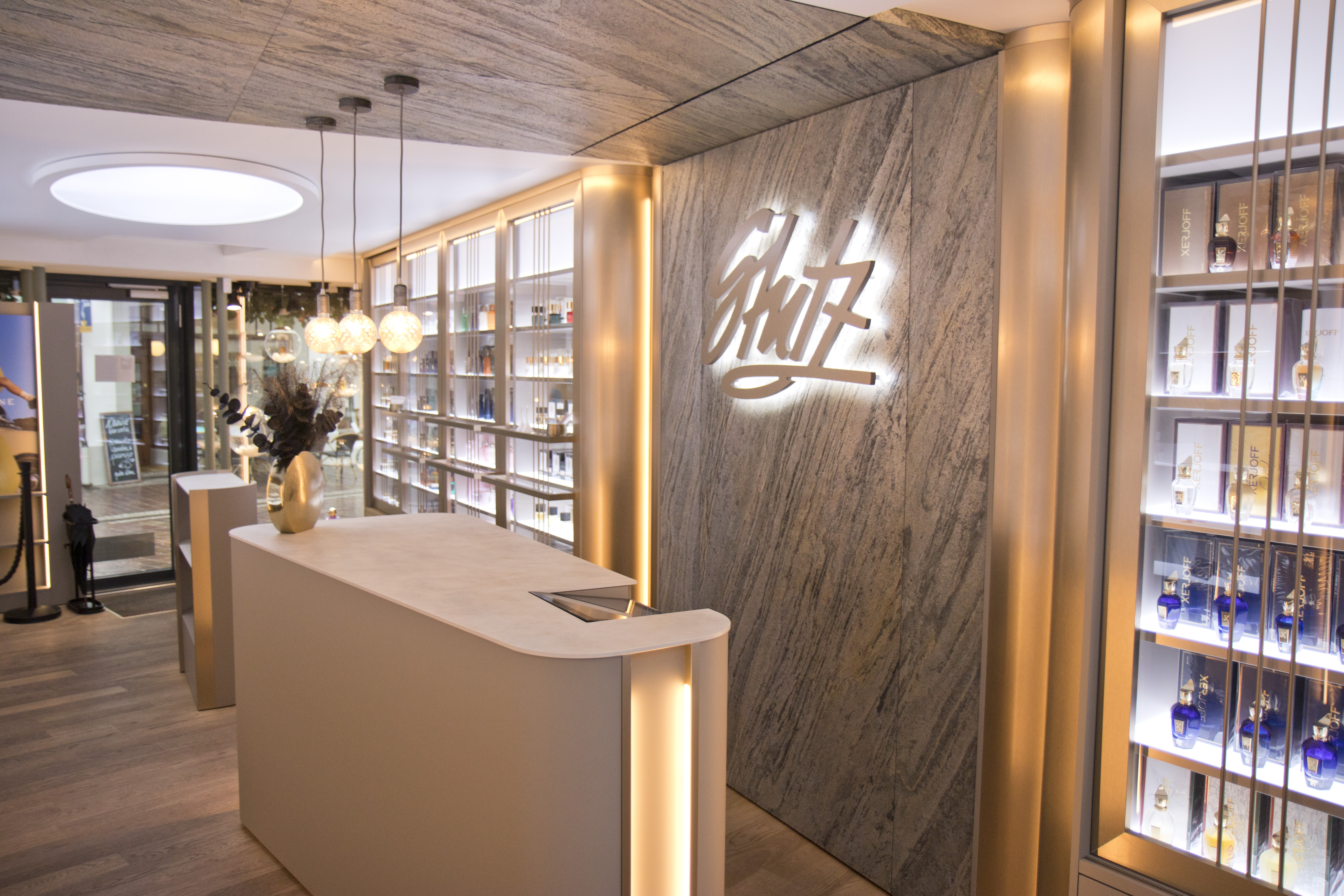 Innenansicht einer eleganten Parfümerie mit modernem Design. Ein heller Empfangstresen mit Blumenvase steht im Vordergrund, dahinter beleuchtete Regale voller Parfumflakons. An der Wand leuchtet das stilisierte Logo „Schütz“ in weißer Schrift auf marmoriertem Hintergrund, warme Lichtakzente sorgen für eine einladende Atmosphäre.