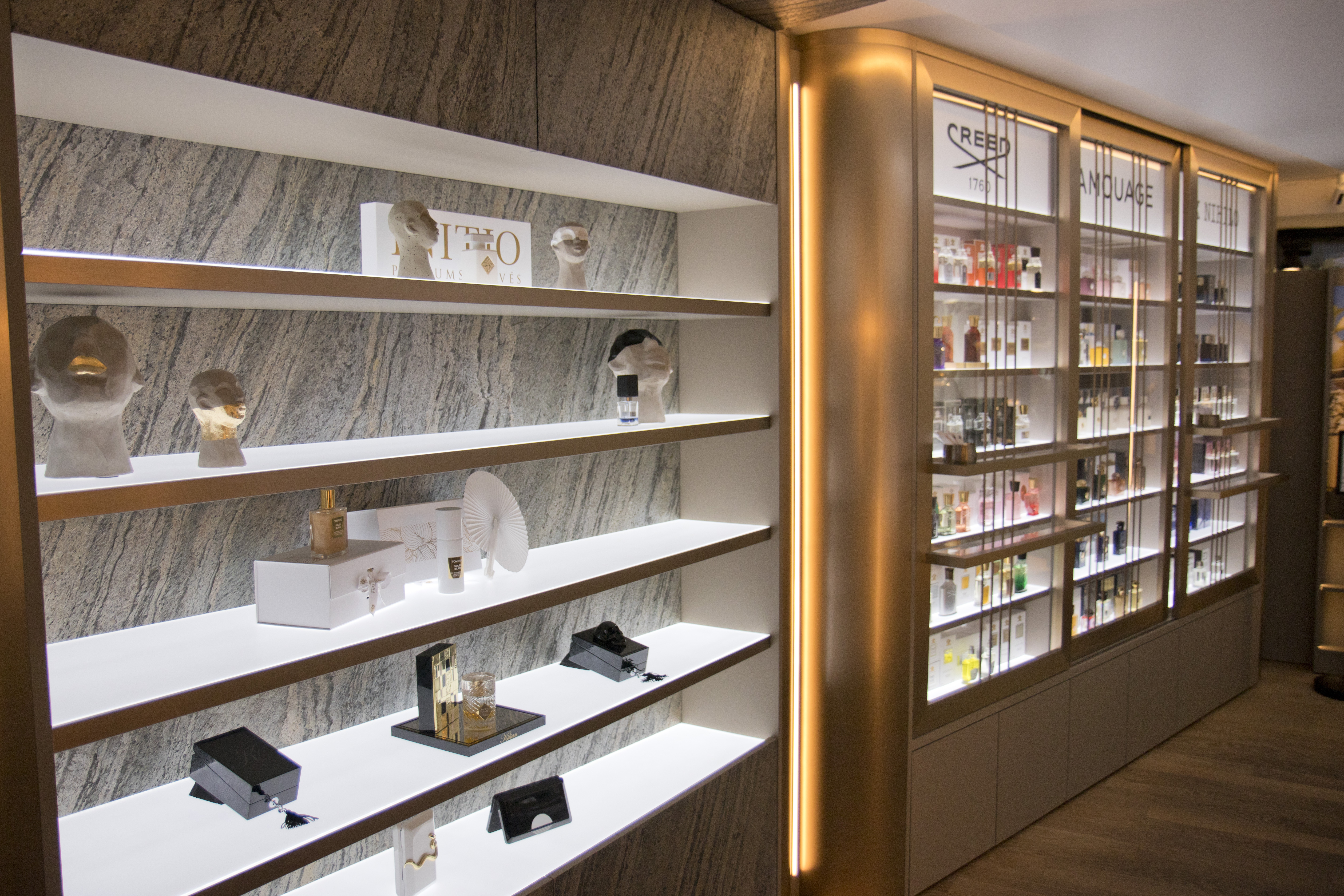 Innenaufnahme einer luxuriösen Parfümerie mit beleuchteten Präsentationsregalen. Links sind auf weißen Regalböden künstlerische Skulpturen und Designerparfums ausgestellt, darunter Düfte der Marke Initio. Rechts im Bild stehen beleuchtete Glasregale mit edlen Parfums von Marken wie Creed und Amouage. Die warme Beleuchtung und goldfarbenen Akzente schaffen eine exklusive Atmosphäre.