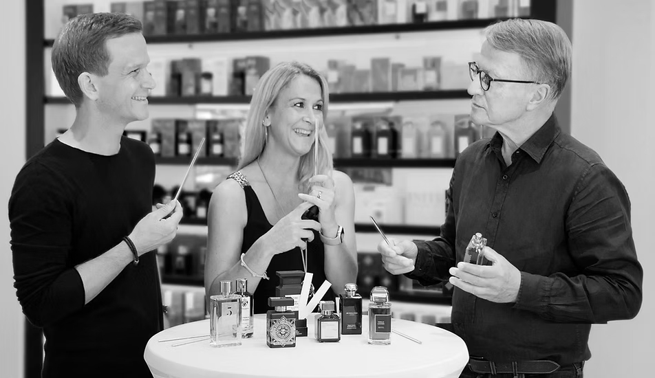 Drei Personen stehen lächelnd an einem Tisch in einer modernen Parfümerie, probieren verschiedene Düfte und besprechen Parfümflakons, die vor ihnen stehen.