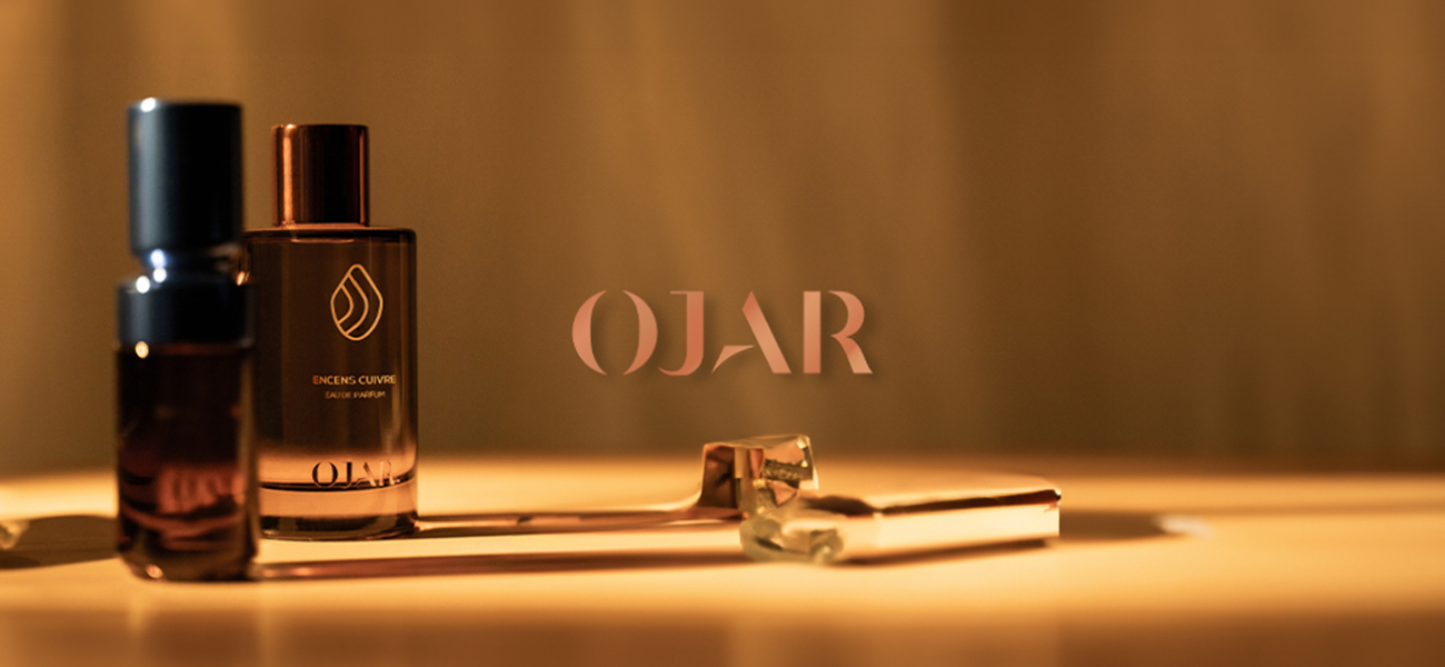 Stimmungsvolles Werbebild für die Marke OJAR. Zwei elegante Parfumflakons, einer mit dem Aufdruck 'ENCENS CUIVRÉ', stehen auf einer warm beleuchteten Fläche. Davor liegt ein goldfarbenes Accessoire, möglicherweise ein Applikator. In der Mitte steht der Markenname 'OJAR' in roségoldener Schrift.
