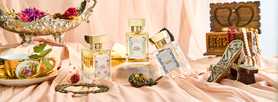 Elegantes Stillleben mit mehreren Regalien Parfumflakons auf roséfarbenem Stoff, umgeben von goldenen Accessoires, Schmuck, Blüten und dekorativen Vintage-Elementen.