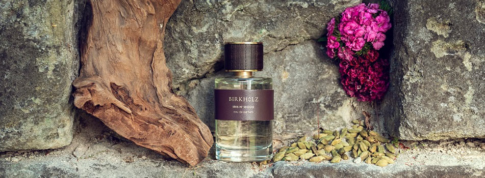 Parfumflakon „Iris N’ Wood“ von Birkholz steht auf einem Steinuntergrund neben einem Stück Treibholz, grünem Kardamom und pinken Blüten.
