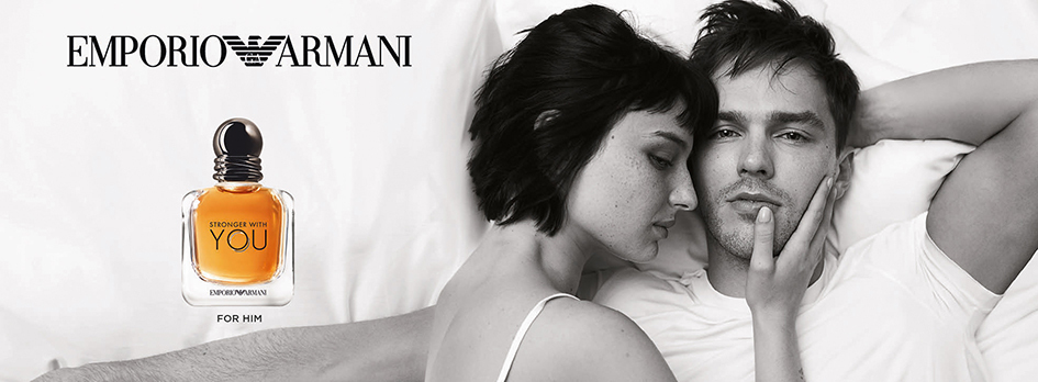 Werbebild für 'Emporio Armani Stronger With You – For Him'. Rechts liegt ein Paar in einem Bett, in einer intimen Schwarz-Weiß-Aufnahme. Die Frau lehnt sich an den Mann, der ernst in die Kamera blickt. Links auf dem weißen Kissen ist der Parfumflakon mit bernsteinfarbener Flüssigkeit und rundem, dunklem Verschluss zu sehen. Darüber steht das Logo 'Emporio Armani'.