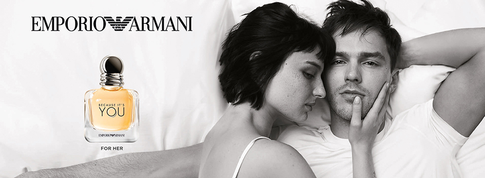 Werbebild für 'Emporio Armani Because It’s You – For Her'. Rechts liegt ein verliebtes Paar in schwarz-weiß auf einem Bett, die Frau lehnt sich zärtlich an den Mann. Links ist der Parfumflakon abgebildet: durchsichtig mit goldenem Duftinhalt und runder silberner Kappe. Darüber steht das Logo 'Emporio Armani'.