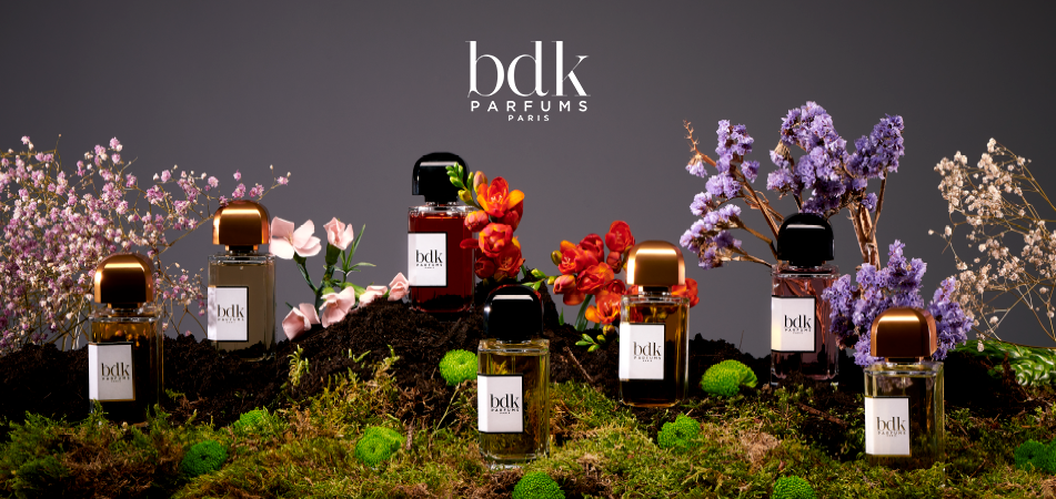 Mehrere Parfumflakons von BDK Parfums Paris stehen auf moosbedecktem Untergrund, umgeben von bunten Blüten in Violett, Rosa, Rot und Weiß. Darüber der Schriftzug „bdk Parfums Paris“ auf dunklem Hintergrund.