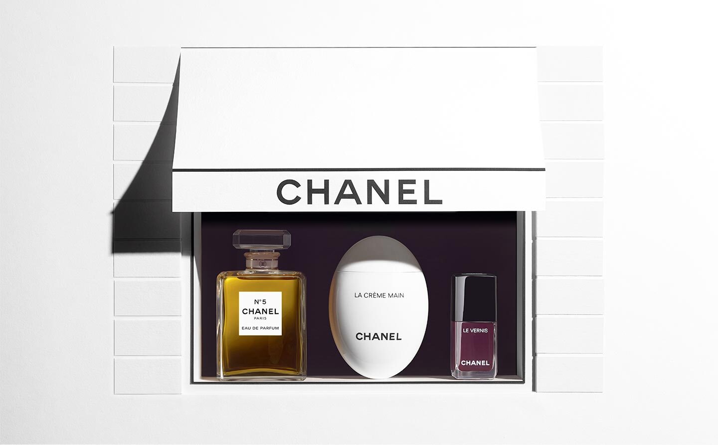 Abgebildet sind drei Chanel-Produkte in einer stilisierten Schaufensterdarstellung mit weißem Baldachin und dem Schriftzug „CHANEL“: links das Parfum Chanel N°5 Eau de Parfum in einem rechteckigen Glasflakon, in der Mitte die weiße Handcreme „La Crème Main“ in ovaler Form, rechts ein dunkelroter Nagellack „Le Vernis“.