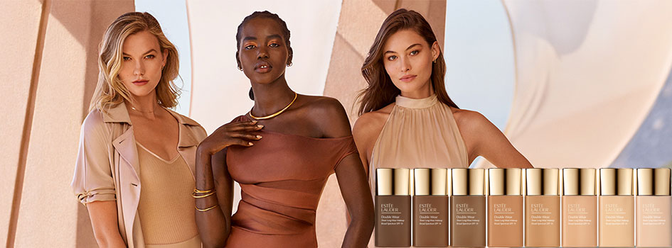 Drei Models mit unterschiedlichen Hauttönen in eleganter, beiger Kleidung posieren vor einem warmen, textilen Hintergrund. Rechts im Bild stehen mehrere Foundation-Produkte von Estée Lauder in verschiedenen Farbtönen.