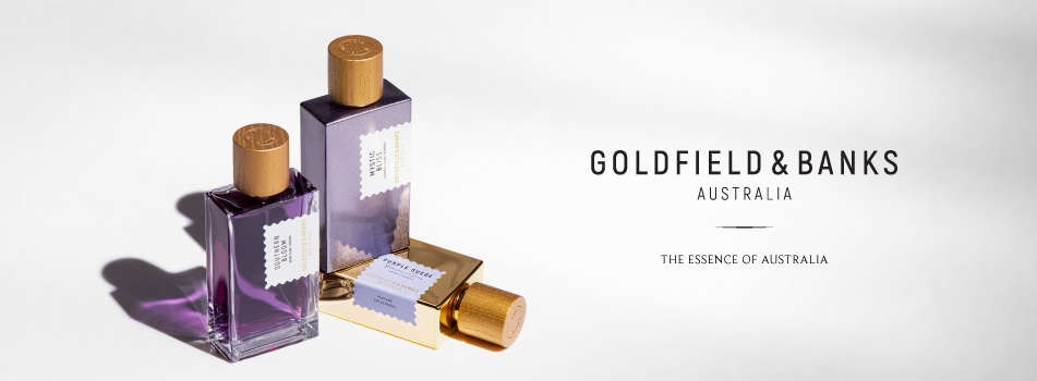 Werbebild für Goldfield & Banks Australia. Zwei stehende und ein liegender Parfumflakon mit violettem Inhalt und goldener Kappe sind auf einer hellen Fläche arrangiert. Die Flakons tragen weiße Etiketten mit goldener Umrandung. Rechts im Bild steht in schwarzer Schrift 'Goldfield & Banks Australia' und darunter der Slogan 'The Essence of Australia'.
