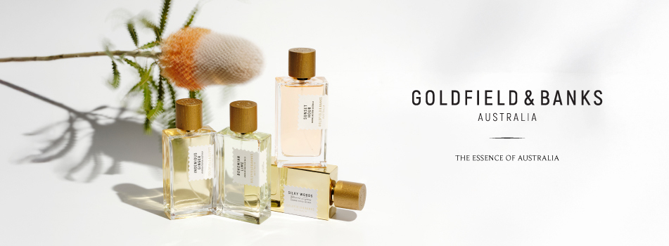 Werbebild für Goldfield & Banks Australia. Vier eckige Parfumflakons mit goldenen Kappen stehen und liegen auf einer weißen Fläche, im Hintergrund ein australischer Banksia-Zweig. Rechts im Bild steht der Markenname 'Goldfield & Banks Australia' und der Slogan 'The Essence of Australia' in schwarzer Schrift.