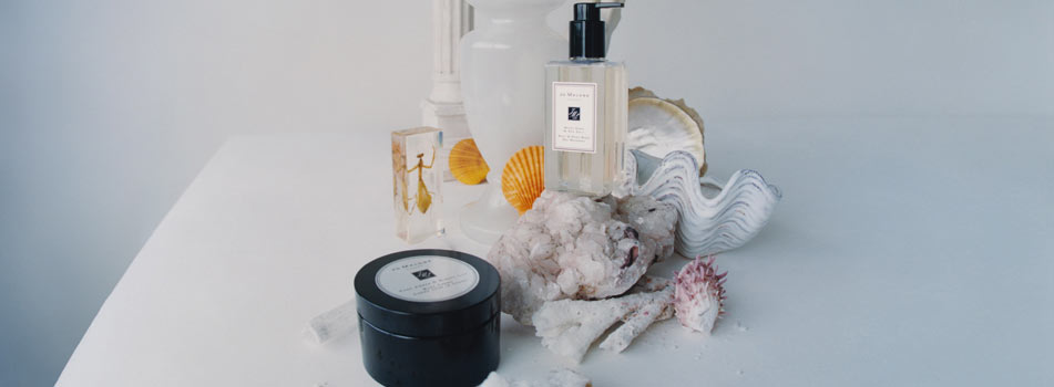 Pflegeprodukte von Jo Malone London sind auf einer weißen Fläche mit Muscheln, Steinen und Dekoration in Strandoptik arrangiert. Zu sehen sind ein Body & Hand Wash in einer Pumpflasche und eine schwarze Dose Body Crème.