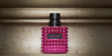 Pinkfarbener Valentino-Parfümflakon mit Nietenstruktur und schwarzem Verschluss steht vor einer marmorierten Steinwand. Auf dem Flakon steht in einem schwarzen Etikett das Wort 'Valentino' in pinker Schrift.