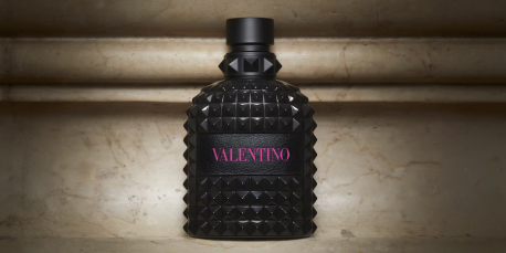 Schwarzer Valentino-Parfümflakon mit Nietenstruktur und schwarzem Verschluss steht vor einer marmorierten Steinwand. Auf dem Flakon steht in einem schwarzen Etikett das Wort 'Valentino' in pinker Schrift.