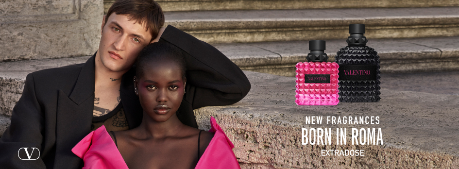 Werbeanzeige für Valentino-Parfüms 'Born in Roma'. Ein schwarzer und ein pinkfarbener Parfümflakon stehen auf einer Steintreppe. Links im Bild posieren ein junger Mann im schwarzen Blazer mit freiem Oberkörper und eine Frau mit dunkler Haut und pinkem, schulterfreiem Kleid. Text auf dem Bild: 'New Fragrances Born in Roma. Extrabose'. Unten links Valentino-Logo.